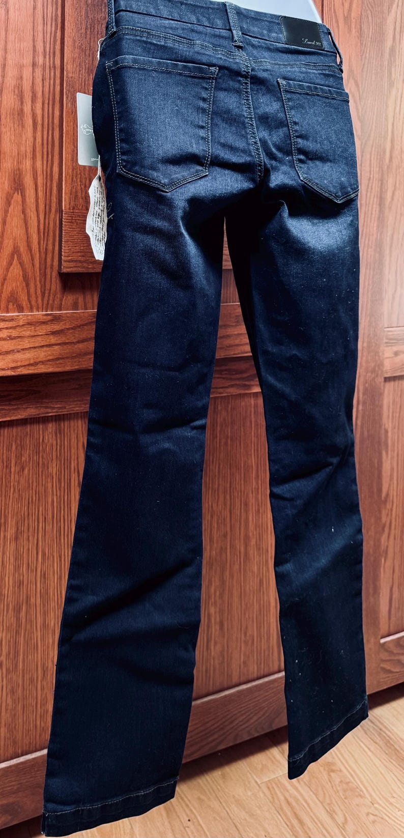 May include: Dark wash denim jeans with a classic five-pocket design. The jeans have a straight leg and a slight fade. A tag is attached to the jeans. The jeans are displayed against a wooden background.