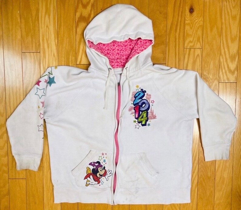 K&ouml;nnte beinhalten: Wei&szlig;er Kapuzenpullover mit Rei&szlig;verschluss und rosa Futter. Der Hoodie hat ein Mickey Mouse-Motiv auf der Vorderseite und ein 2014-Motiv auf der R&uuml;ckseite. Der Hoodie hat Sterne auf den &Auml;rmeln.