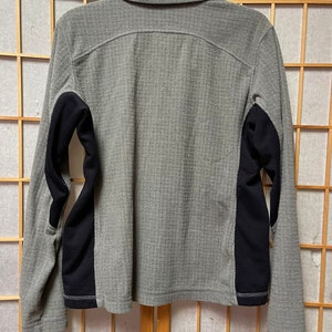 May include: A gray long-sleeved shirt with black panels on the sleeves. The shirt has a button-up collar and a zipper closure.