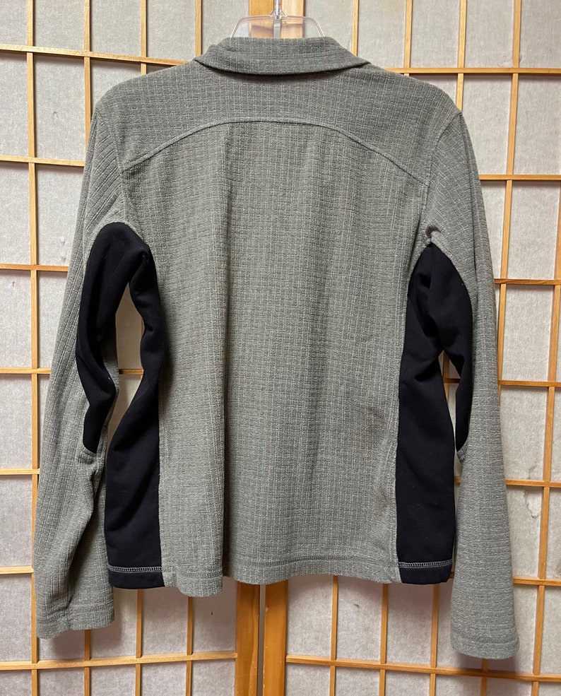 May include: A gray long-sleeved shirt with black panels on the sleeves. The shirt has a button-up collar and a zipper closure.