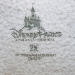 K&ouml;nnte beinhalten: Ein wei&szlig;es Etikett mit grauem Text und einem grauen Bild des Disney-Schlosses. Der Text lautet "Disney Parks Authentic Original 2X RN 129855 Made in Thailand &copy;Disney".