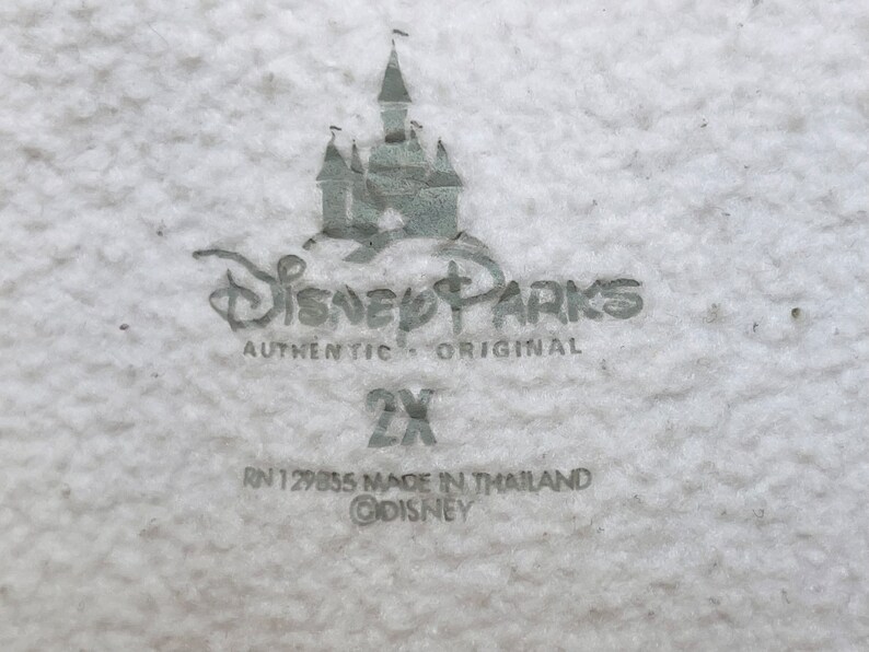 K&ouml;nnte beinhalten: Ein wei&szlig;es Etikett mit grauem Text und einem grauen Bild des Disney-Schlosses. Der Text lautet "Disney Parks Authentic Original 2X RN 129855 Made in Thailand &copy;Disney".