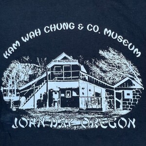 Puede incluir: Impresión gráfica en blanco y negro de un edificio con el texto "KAM WAH CHUNG & CO. MUSEUM" y "JOHN DAY, OREGON" en una camiseta negra.
