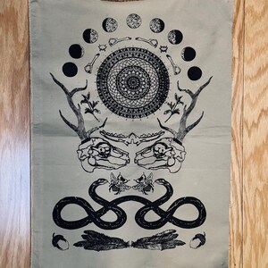 Peut inclure: Une tapisserie en toile beige avec un motif noir et blanc. Le motif représente un mandala, un serpent, deux abeilles, des bois de cerf, un crâne et un croissant de lune.
