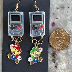 May include: A pair of gold-tone earrings featuring a clear plastic game console charm with red buttons and a gold chain attached to a cartoon character charm. One charm is of Mario, the other is of Luigi.