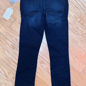 May include: Dark wash denim jeans with a classic five-pocket design. The jeans have a straight leg cut and a leather patch at the back waistband. A tag is attached to the jeans.
