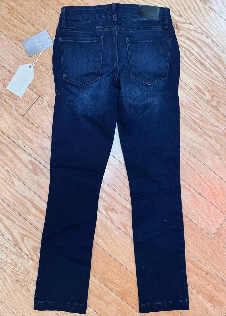 May include: Dark wash denim jeans with a classic five-pocket design. The jeans have a straight leg cut and a leather patch at the back waistband. A tag is attached to the jeans.
