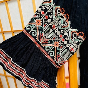 Könnte beinhalten: Schwarzer Stoffärmel mit einem bunten Stickmuster. Die Stickerei zeigt geometrische Muster in Rot-, Grün- und Weißtönen.