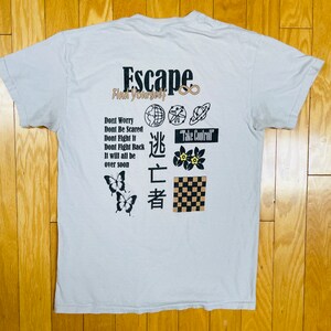 May include: A light grey t-shirt with the text "Escape Find Yourself" in black and brown lettering. The shirt also features a variety of graphics, including butterflies, flowers, a chequered pattern, and the text "逃亡者" in black. The text "Take Control" is also visible in a black rectangle.