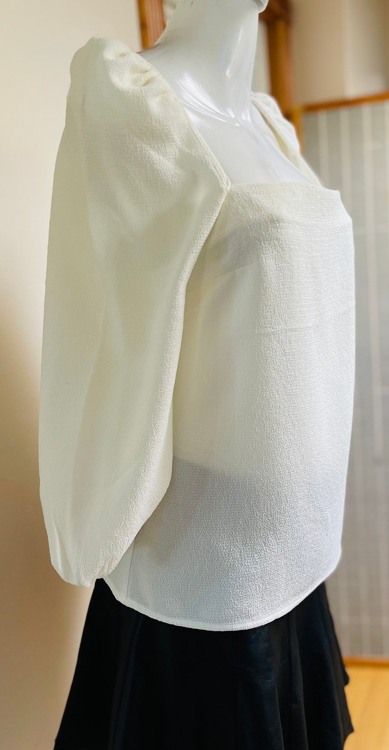 May include: A white, textured,  square-neck blouse with puffed sleeves. The blouse is cropped and has a fitted silhouette.