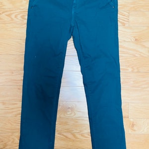 Puede incluir: Pantalones de color verde azulado con cierre de bot&oacute;n y bolsillos. Los pantalones est&aacute;n hechos de una tela lisa y est&aacute;n colocados sobre una superficie de madera. Una etiqueta est&aacute; adherida a la cintura.