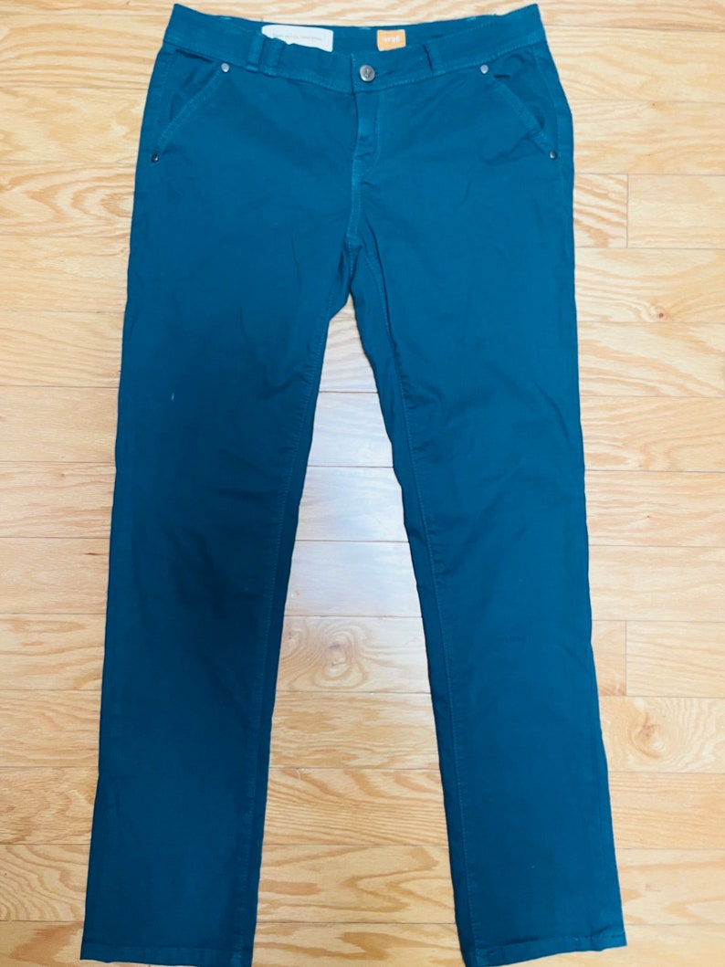 Puede incluir: Pantalones de color verde azulado con cierre de bot&oacute;n y bolsillos. Los pantalones est&aacute;n hechos de una tela lisa y est&aacute;n colocados sobre una superficie de madera. Una etiqueta est&aacute; adherida a la cintura.