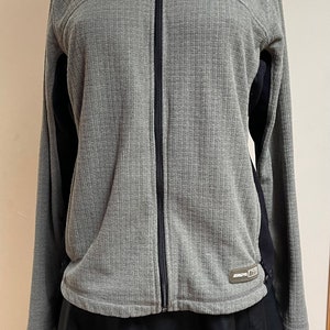 May include: Gray and black zip-up jacket with a ribbed texture and a black logo that says "AGL" on the bottom right side. The jacket is on a mannequin wearing a black skirt.