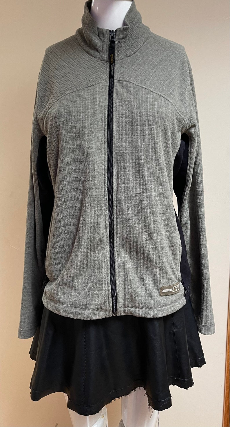 May include: Gray and black zip-up jacket with a ribbed texture and a black logo that says "AGL" on the bottom right side. The jacket is on a mannequin wearing a black skirt.