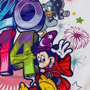 K&ouml;nnte beinhalten: Wei&szlig;er Pullover mit gestickten Bildern von Mickey Mouse als Zauberer, Donald Duck und den Zahlen 2014. Der Hintergrund zeigt Feuerwerk und Sterne.