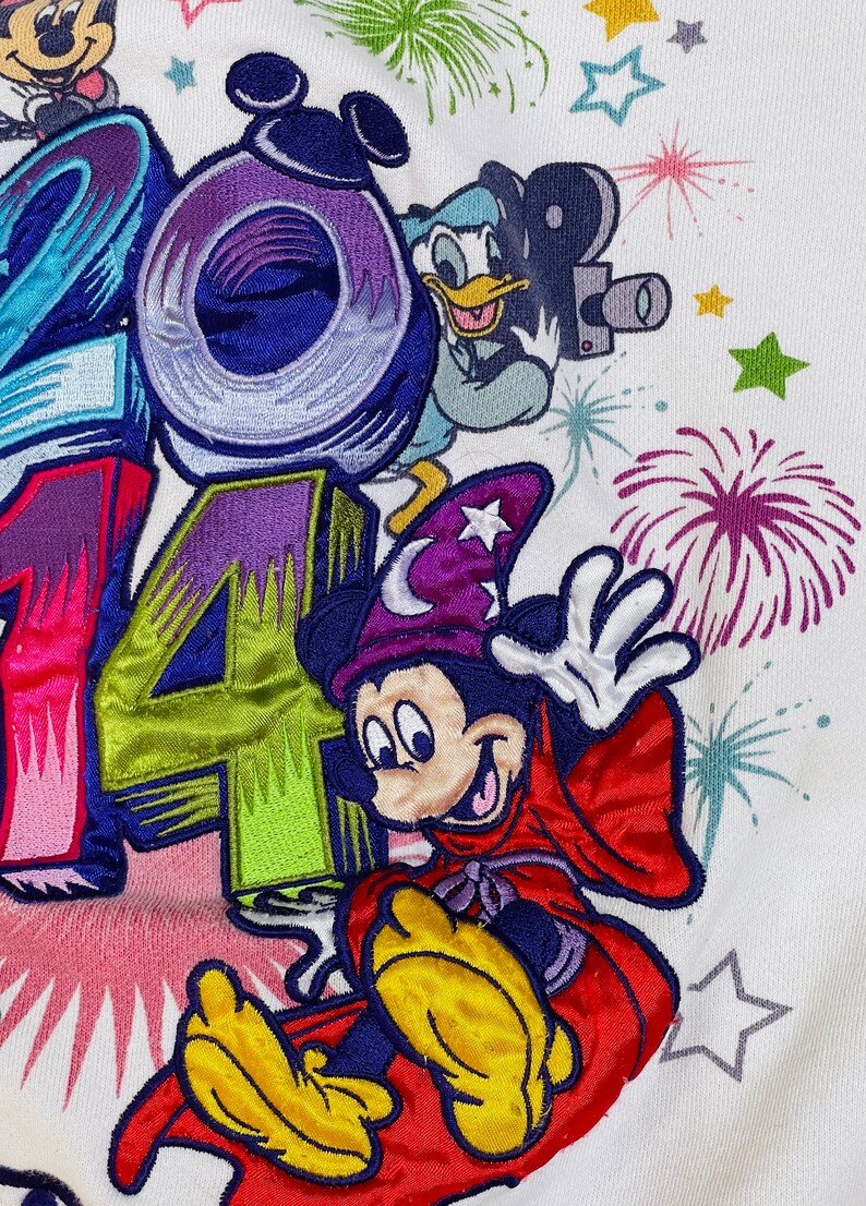 K&ouml;nnte beinhalten: Wei&szlig;er Pullover mit gestickten Bildern von Mickey Mouse als Zauberer, Donald Duck und den Zahlen 2014. Der Hintergrund zeigt Feuerwerk und Sterne.