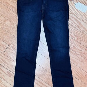 May include: Dark wash denim jeans with a classic five-pocket design. The jeans have a straight leg cut and a button and zip closure. A tag is attached to the jeans. The jeans are displayed on a wooden surface.