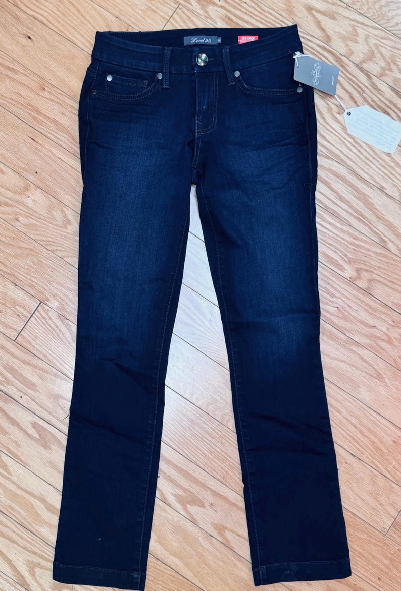 May include: Dark wash denim jeans with a classic five-pocket design. The jeans have a straight leg cut and a button and zip closure. A tag is attached to the jeans. The jeans are displayed on a wooden surface.