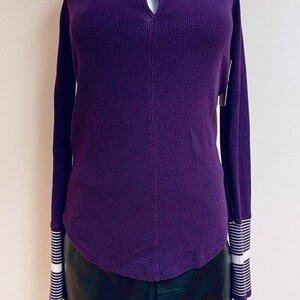 May include: A purple waffle knit long-sleeved shirt with a keyhole neckline and white striped cuffs. The shirt is on a white mannequin and has a relaxed fit.