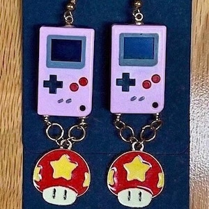 Puede incluir: Un par de pendientes de Game Boy rosa y morado con un amuleto de hongo rojo y amarillo. Los pendientes están hechos de metal y tienen un acabado dorado.