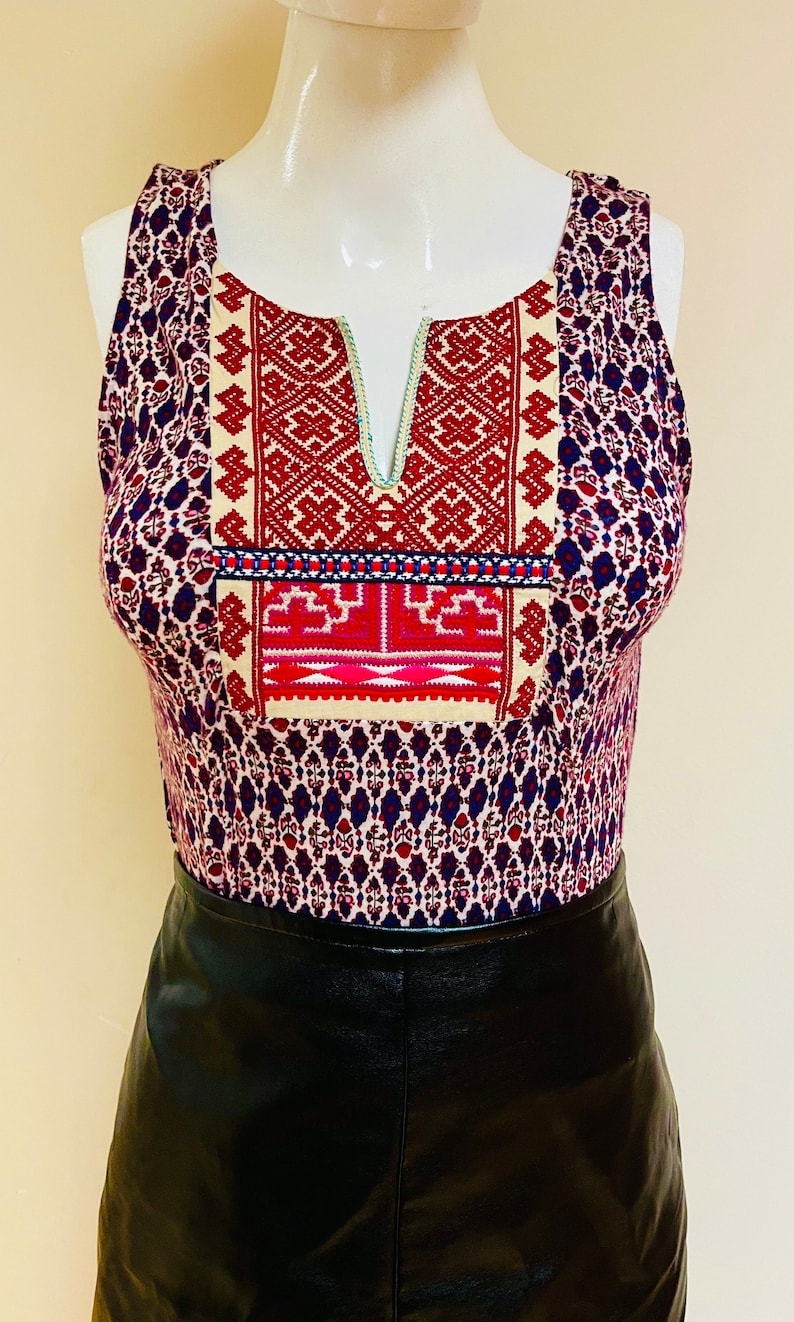 May include: A sleeveless top with a white, red, and blue embroidered design on the front. The top is made of a patterned fabric with a blue, red, and white design. The top is paired with a black leather skirt.