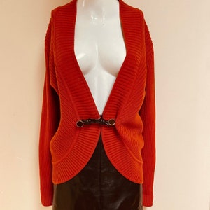 May include: A rust-colored knitted cardigan with a deep V-neck and a long sleeve. The cardigan has a unique closure with two metal rings and a leather strap. The cardigan is worn over a black leather skirt.