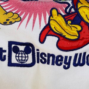 K&ouml;nnte beinhalten: Wei&szlig;es T-Shirt mit einem bunten gestickten Design mit Mickey Mouse und Pluto. Das Shirt hat auch den Text "Walt Disney World" in Blau gestickt.