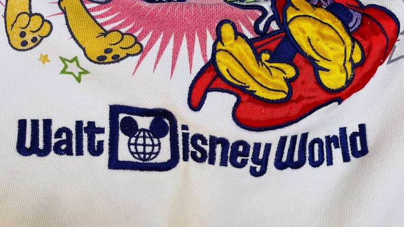 K&ouml;nnte beinhalten: Wei&szlig;es T-Shirt mit einem bunten gestickten Design mit Mickey Mouse und Pluto. Das Shirt hat auch den Text "Walt Disney World" in Blau gestickt.