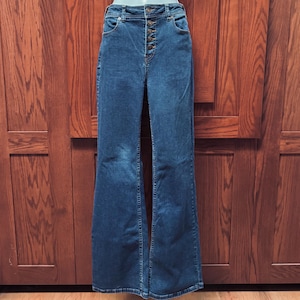 May include: Dark wash denim flare jeans with a high waist and button fly. The jeans have a classic five-pocket design and a wide leg opening. The jeans are displayed on a mannequin against a wooden background.