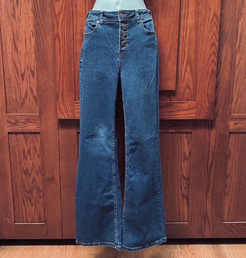 May include: Dark wash denim flare jeans with a high waist and button fly. The jeans have a classic five-pocket design and a wide leg opening. The jeans are displayed on a mannequin against a wooden background.