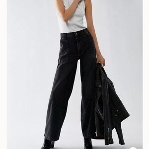 May include: A woman wearing a white tank top and black high-rise jeans, holding a black jacket. The image is from the brand "We The Free". The jeans have large pockets. The model is wearing black boots.