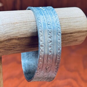Puede incluir: Un brazalete de metal plateado con un patrón geométrico. El brazalete está en un expositor de madera.