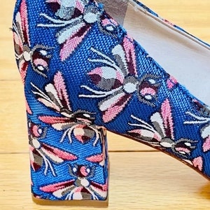 May include: A blue high-heeled shoe with a floral pattern featuring pink and white butterflies. The heel is a block heel and is covered in the same floral pattern.
