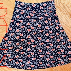 May include: A-line skirt with a navy blue background and a repeating floral pattern in coral and white. The skirt is made of a lightweight fabric and has a comfortable waistband. The skirt is suitable for casual wear.