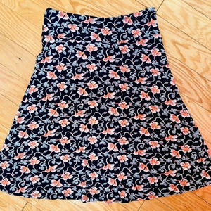 May include: A-line skirt with a floral pattern. The skirt features a dark navy blue background with a repeating pattern of pink flowers and white vines. The skirt is made of a lightweight fabric.