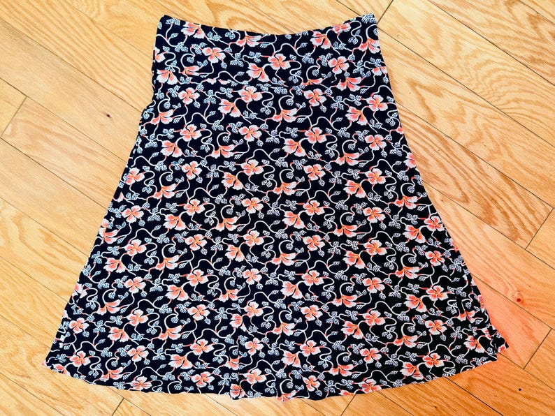 May include: A-line skirt with a floral pattern. The skirt features a dark navy blue background with a repeating pattern of pink flowers and white vines. The skirt is made of a lightweight fabric.