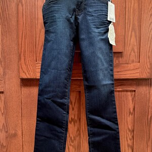 May include: Dark wash denim jeans with a straight leg cut. The jeans feature a classic five-pocket design, a button and zip closure, and a tag attached to the waistband. The jeans are displayed against a wooden door background.