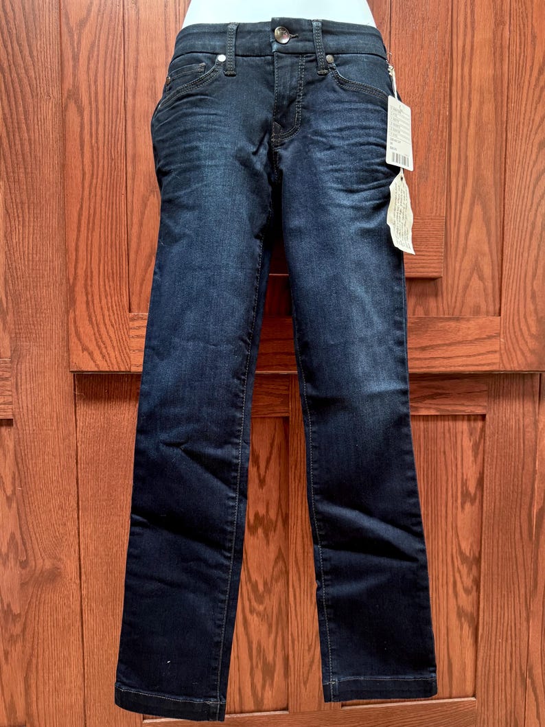May include: Dark wash denim jeans with a straight leg cut. The jeans feature a classic five-pocket design, a button and zip closure, and a tag attached to the waistband. The jeans are displayed against a wooden door background.