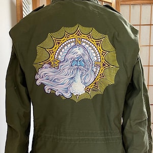 May include: Olive green military-style jacket with a zipper closure and a unique graphic on the back. The graphic features a bearded, blue-skinned figure blowing a cloud of white vapor, surrounded by a yellow and purple sunburst design.