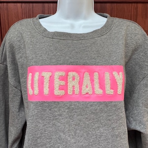 Op de afbeelding: Een grijze sweatshirt met het woord "LITERALLY" in witte, getextureerde letters op een felroze rechthoekige patch over de borst. De sweatshirt heeft een ronde hals en lange mouwen.