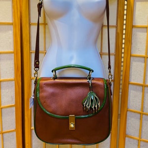 May include: A brown leather satchel bag with a green trim and a gold-toned clasp. The bag has a detachable shoulder strap and a small, decorative tassel.