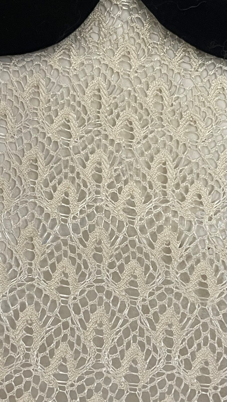 May include: A close-up of a white crocheted fabric with a delicate, intricate pattern. The fabric has a soft, airy texture and a subtle sheen.
