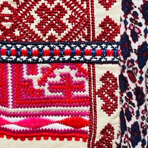 May include: Close-up of a white fabric with red, pink, and blue embroidery. The embroidery features geometric patterns and floral motifs. The fabric is part of a larger piece of clothing or textile.