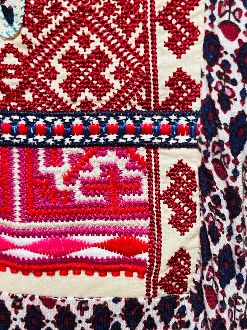 May include: Close-up of a white fabric with red, pink, and blue embroidery. The embroidery features geometric patterns and floral motifs. The fabric is part of a larger piece of clothing or textile.