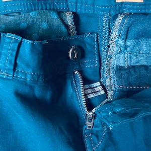 Puede incluir: Primer plano de unos pantalones de color verde azulado con una cremallera parcialmente desabrochada. Los pantalones tienen un cierre de bot&oacute;n y una etiqueta que dice "Pilcro and the Letterpress". Otra etiqueta muestra "No 26."