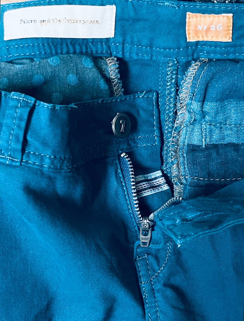 Puede incluir: Primer plano de unos pantalones de color verde azulado con una cremallera parcialmente desabrochada. Los pantalones tienen un cierre de bot&oacute;n y una etiqueta que dice "Pilcro and the Letterpress". Otra etiqueta muestra "No 26."