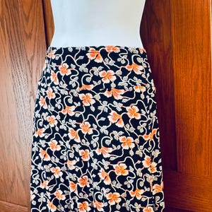 May include: A black skirt with a floral pattern in orange and white. The skirt is displayed on a mannequin. The floral design features orange flowers and white leaves on a black background. The skirt appears to be knee-length.