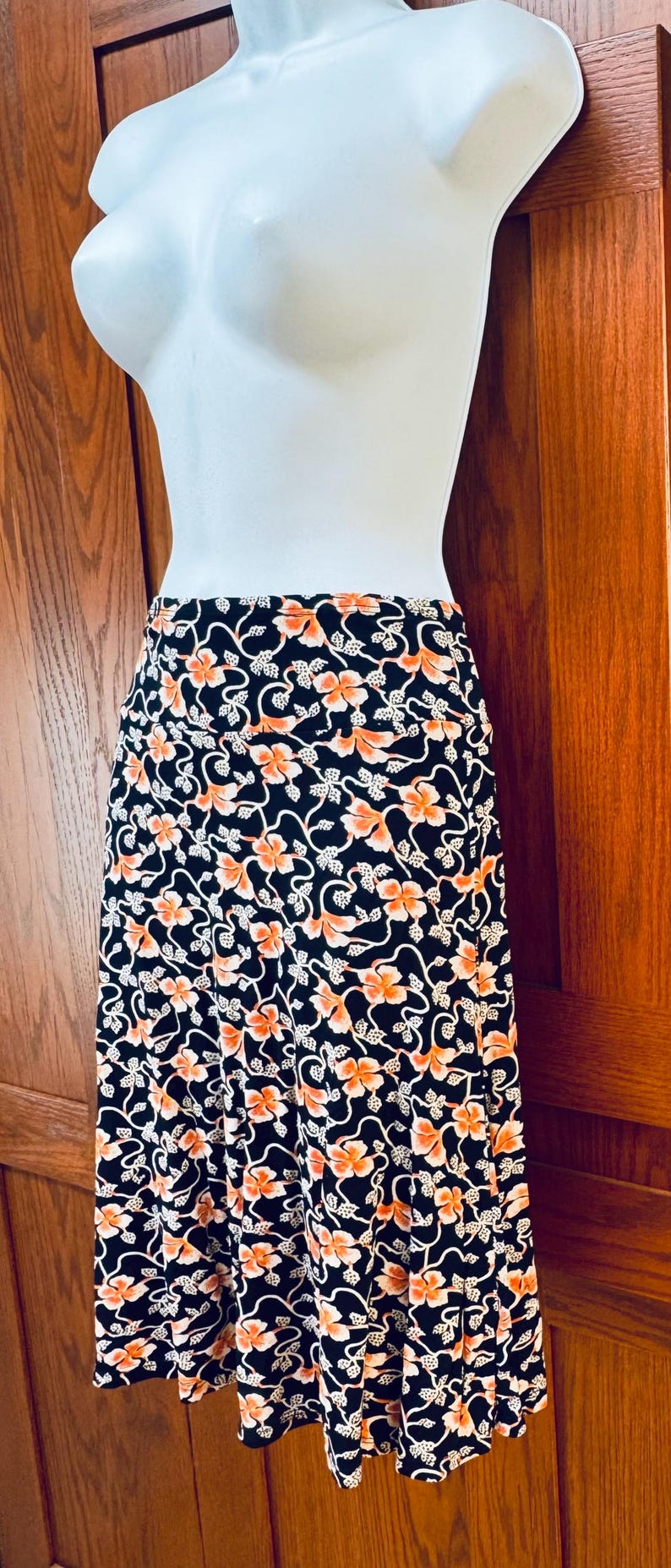 May include: A black skirt with a floral pattern in orange and white. The skirt is displayed on a mannequin. The floral design features orange flowers and white leaves on a black background. The skirt appears to be knee-length.