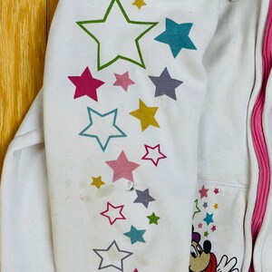 K&ouml;nnte beinhalten: Wei&szlig;er Kapuzenpullover mit rosa Rei&szlig;verschluss und einem Mickey Mouse-Motiv auf dem &Auml;rmel. Der &Auml;rmel hat ein Muster aus bunten Sternen.