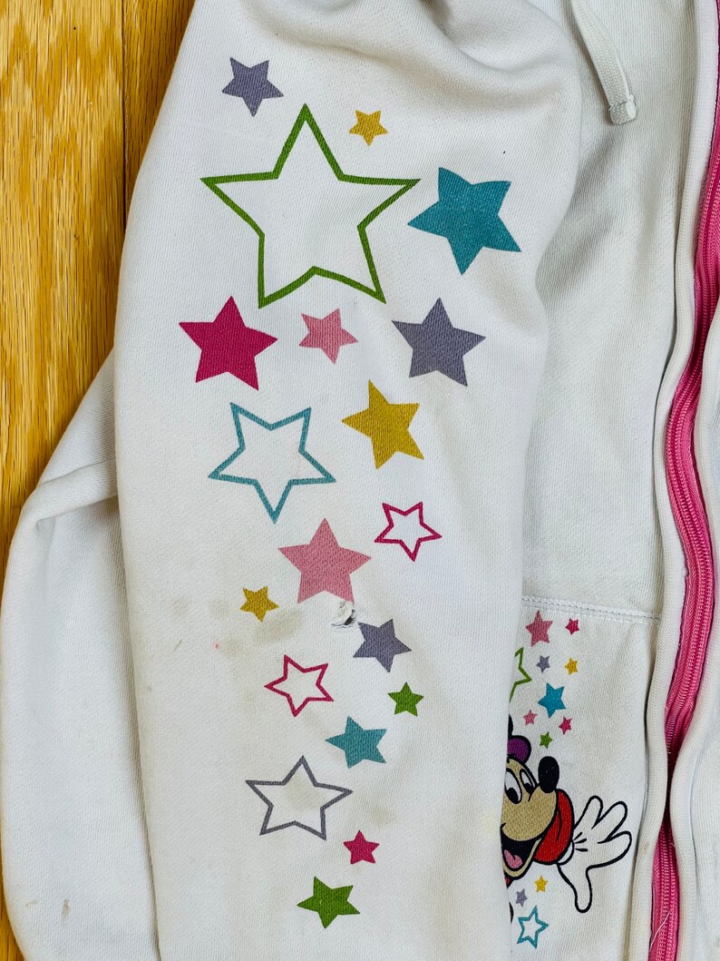 K&ouml;nnte beinhalten: Wei&szlig;er Kapuzenpullover mit rosa Rei&szlig;verschluss und einem Mickey Mouse-Motiv auf dem &Auml;rmel. Der &Auml;rmel hat ein Muster aus bunten Sternen.